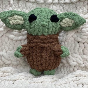 May include: A handmade crochet doll in the likeness of a popular character. The doll features large, pointed ears, black eyes, and a green head and hands. The doll's body is brown, and it is set against a white, textured background.