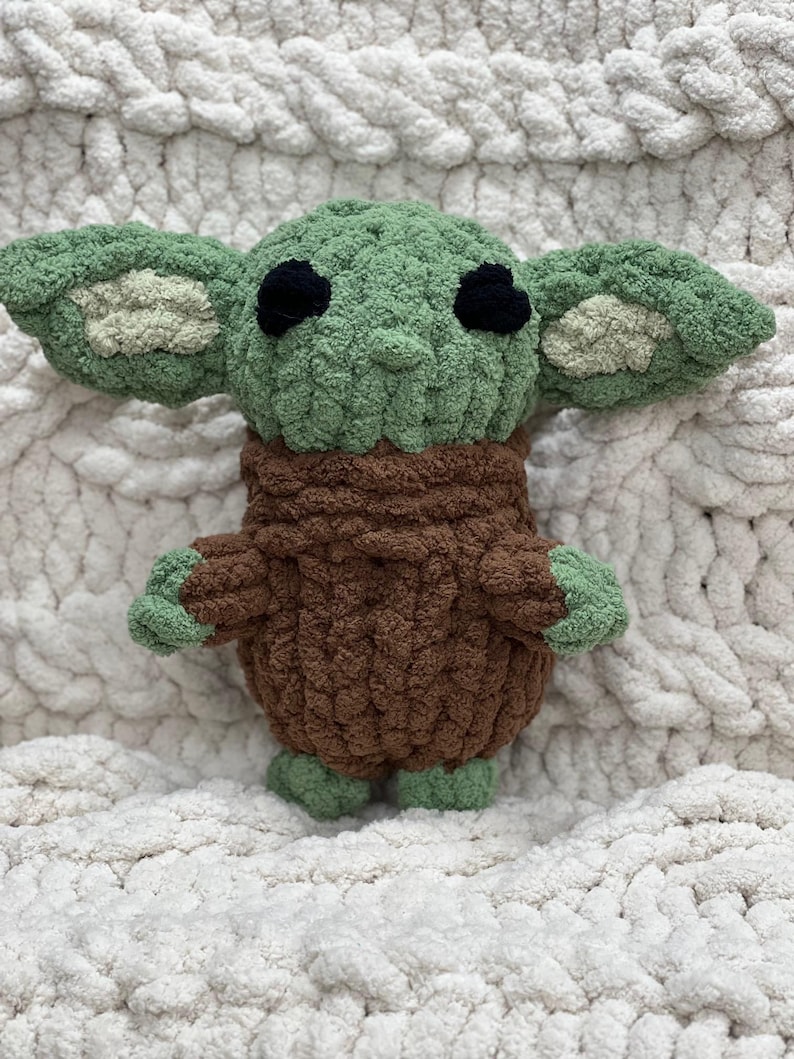 May include: A handmade crochet doll in the likeness of a popular character. The doll features large, pointed ears, black eyes, and a green head and hands. The doll's body is brown, and it is set against a white, textured background.