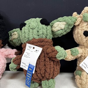 May include: A handmade, green and brown crocheted doll with large ears and a brown robe. The doll has black eyes and a blue ribbon that says "First" and "Marion County Fair". The doll is displayed with other crocheted dolls.
