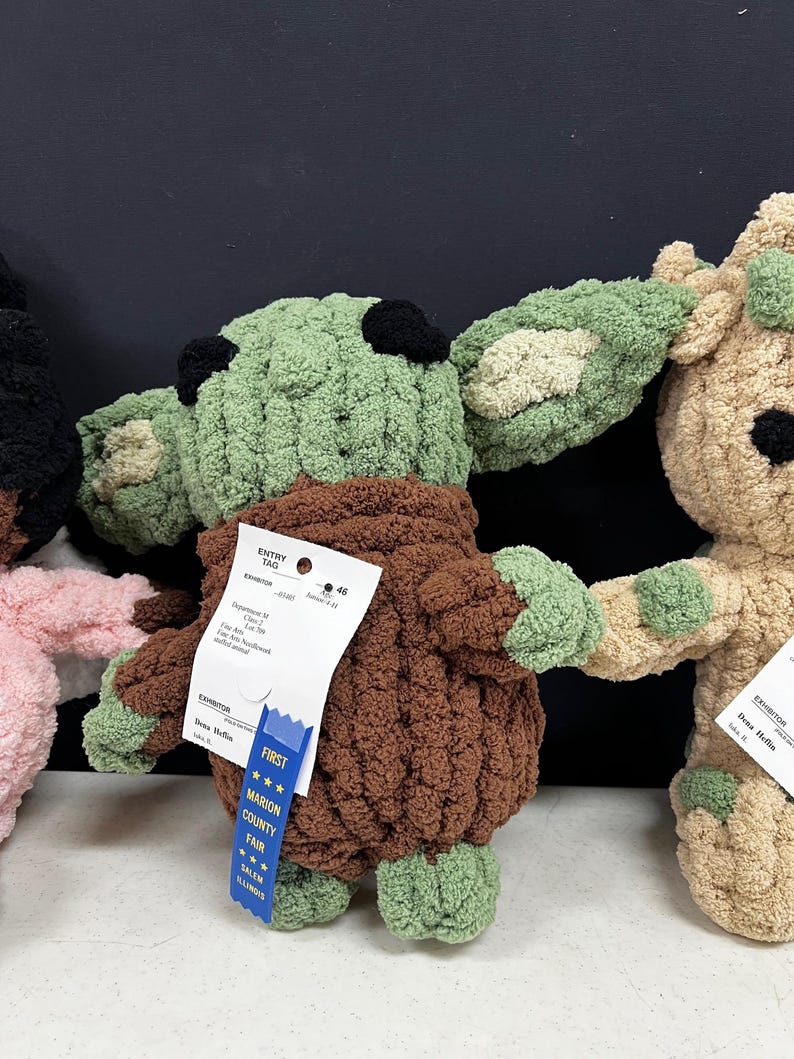 May include: A handmade, green and brown crocheted doll with large ears and a brown robe. The doll has black eyes and a blue ribbon that says "First" and "Marion County Fair". The doll is displayed with other crocheted dolls.