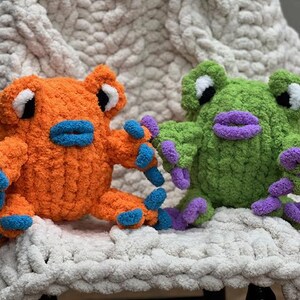 May include: Two crocheted frog plushies, one orange with blue accents and one green with purple accents. The plushies are sitting on a white fluffy blanket.