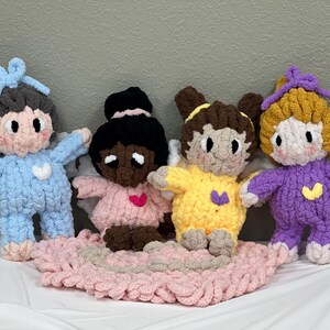 K&ouml;nnte beinhalten: Vier geh&auml;kelte Engelpuppen mit Fl&uuml;geln. Die Puppen tragen verschiedene farbige Outfits: blau, rosa, gelb und lila. Die Puppen sitzen auf einer rosa geh&auml;kelten Decke.