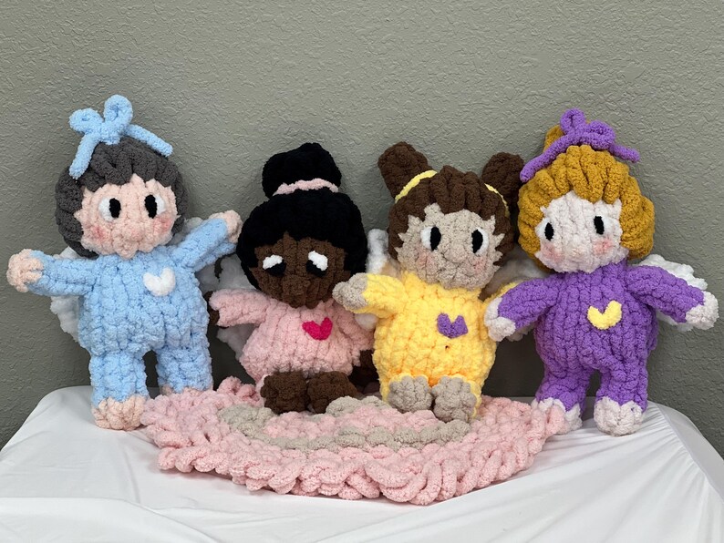 K&ouml;nnte beinhalten: Vier geh&auml;kelte Engelpuppen mit Fl&uuml;geln. Die Puppen tragen verschiedene farbige Outfits: blau, rosa, gelb und lila. Die Puppen sitzen auf einer rosa geh&auml;kelten Decke.