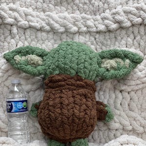 May include: A handmade green and brown crochet doll with large ears, resembling a popular character. The doll is placed on a white, chunky knit blanket. A clear plastic water bottle is next to the doll.
