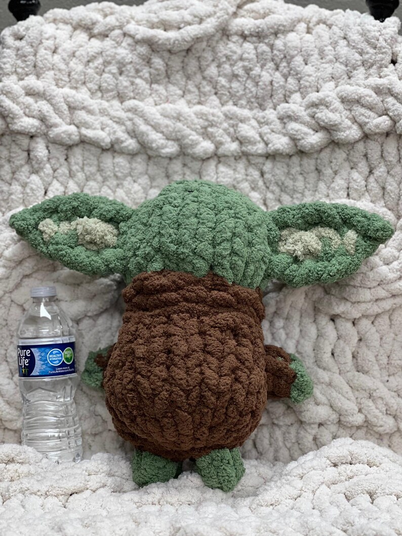 May include: A handmade green and brown crochet doll with large ears, resembling a popular character. The doll is placed on a white, chunky knit blanket. A clear plastic water bottle is next to the doll.