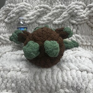 May include: A handmade, brown and green crocheted turtle toy. The turtle has a rounded body, green flippers, and a small head. The toy is placed on a white, chunky knit blanket.