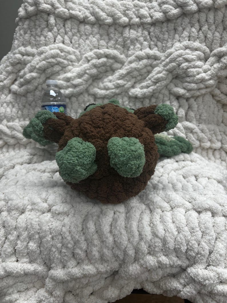 May include: A handmade, brown and green crocheted turtle toy. The turtle has a rounded body, green flippers, and a small head. The toy is placed on a white, chunky knit blanket.