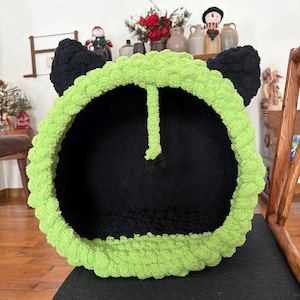 Puede incluir: Una cama para gatos hecha a mano con forma de cabeza de gato. El interior es negro, con un borde verde brillante y orejas negras. La cama está hecha de un hilo grueso y suave y está colocada sobre una superficie negra.