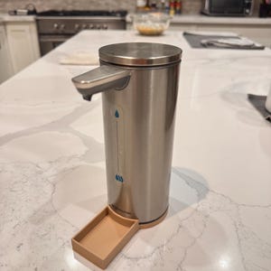 May include: A stainless steel automatic soap dispenser with a beige plastic drip tray. The dispenser has a blue drop icon and the word "simplehuman" printed on the bottom.