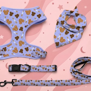 May include: A light purple pet harness, collar, leash, and bandana set featuring a repeating pattern of brown bear-shaped treats and white sticks. The harness has black plastic buckles, the collar has a black buckle, and the leash has a black metal clip.