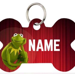 Peut inclure: Une étiquette pour animaux de compagnie en forme d'os rouge avec le mot "NAME" en blanc. L'étiquette présente un personnage de grenouille verte de dessin animé sur le côté gauche. Un anneau en métal argenté est attaché en haut.
