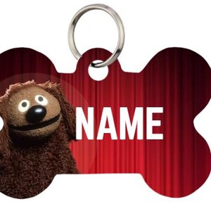 May include: Bone-shaped pet tag with a red background and the word "NAME" in white. A brown puppet dog with a black nose and white eyes is on the left side of the tag. A silver ring is at the top.