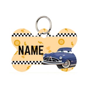 May include: Bone-shaped pet tag with a navy blue cartoon car graphic. The tag has a yellow background with lightning bolts, wheels, and a trophy. The word "NAME" is printed in bold black letters. A silver ring is attached for easy attachment to a pet's collar.