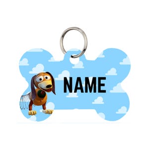 Può includere: Un'etichetta per animali a forma di osso di cane con un'immagine a cartoni animati di Slinky Dog di Toy Story. L'etichetta ha uno sfondo azzurro con nuvole bianche e la parola "NAME" in nero. È presente un anello in metallo argentato.