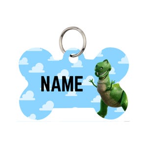 Pode incluir: Uma etiqueta para animais de estimação em forma de osso com um fundo de céu azul e nuvens. A etiqueta apresenta a palavra "NAME" em letras pretas em negrito e um personagem de dinossauro verde. Um anel de metal prateado está preso no topo.
