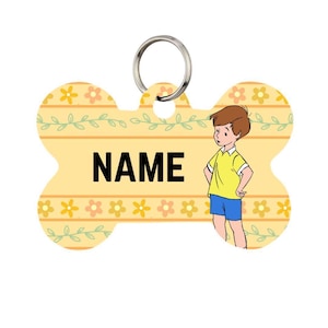Op de afbeelding: Een gele, botvormige hondenpenning met het woord "NAME" in het zwart. De penning heeft een cartoonillustratie van een jongen in een geel shirt en een blauwe korte broek. De penning heeft een zilveren ring voor bevestiging en een bloemenpatroon.