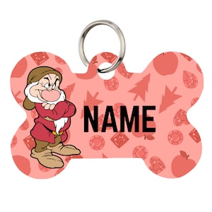 May include: A bone-shaped pet ID tag with a silver metal ring. The tag features a light pink background with a repeating pattern of red apples, pine trees, and gemstones. On the left, a cartoon character with a brown hat and red tunic stands with arms crossed. The word NAME is printed in bold black capital letters in the center.