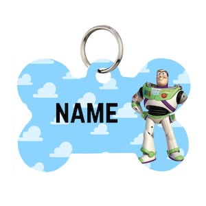 Puede incluir: Una placa para mascotas en forma de hueso, azul claro, con Buzz Lightyear. El fondo es azul cielo con nubes blancas y la palabra "NAME". Un anillo de metal plateado en la parte superior.
