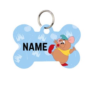 May include: A light blue, bone-shaped pet tag with a silver ring. The tag features a cartoon mouse character in a yellow shirt and red shoes. The word "NAME" is printed in black. The background has a pattern of castles and pumpkins.