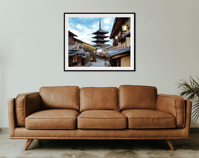 May include: A photograph of a traditional Japanese pagoda in a narrow street lined with buildings. The pagoda is a five-storey structure with a distinctive curved roof. The street is lined with traditional Japanese buildings with wooden facades and tiled roofs. The image is taken from a low angle, looking up at the pagoda.