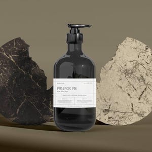 Puede incluir: Una botella negra con bomba de jabón de manos con aroma a tarta de calabaza con una etiqueta blanca. La botella está flanqueada por dos formas de piedra abstractas y agrietadas en marrón oscuro y beige.