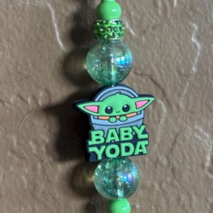 May include: Green and black charm with a cartoon image of Baby Yoda, the text "BABY YODA", and green and iridescent beads.