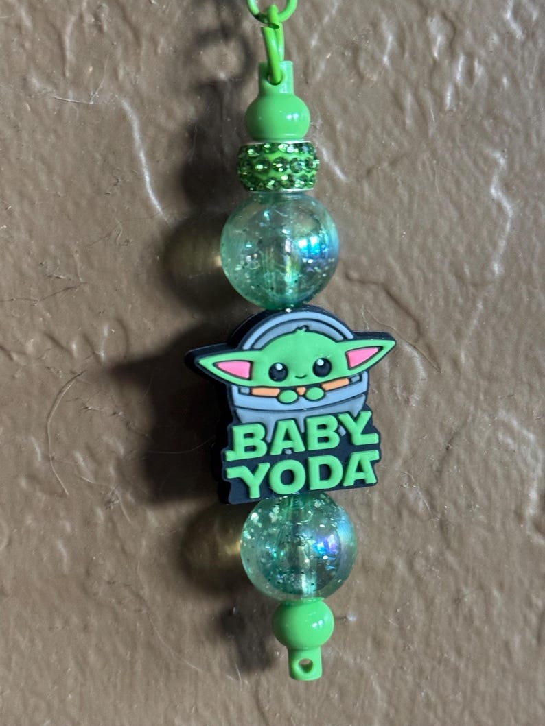 May include: Green and black charm with a cartoon image of Baby Yoda, the text "BABY YODA", and green and iridescent beads.