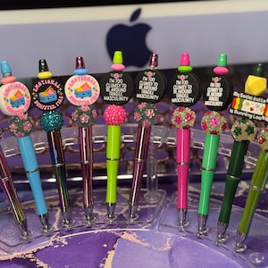 May include: A set of eight colorful pens with different designs and sayings. The pens have a variety of colorful beads and charms. The sayings on the pens include "Emotional Dumpster Fire", "I'm Too Clumsy To Be Around Fragile Masculinity", and "My Social Battery Is Running Low, Like, Low."