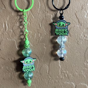 May include: Two green and black Baby Yoda keychains with clear and green glitter beads. The keychains have a green ring and a black ring.