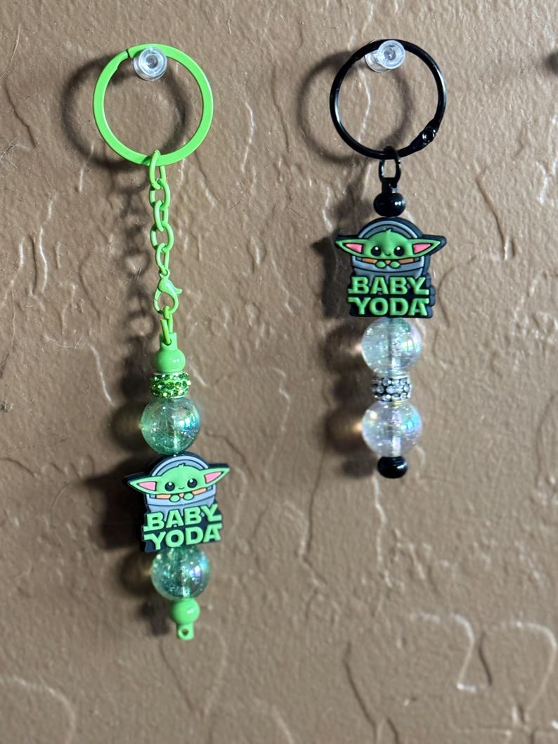 May include: Two green and black Baby Yoda keychains with clear and green glitter beads. The keychains have a green ring and a black ring.