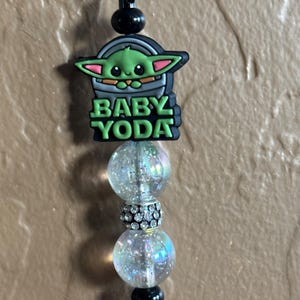 May include: A black and green charm with a cartoon image of Baby Yoda, the words "Baby Yoda" and three iridescent beads.