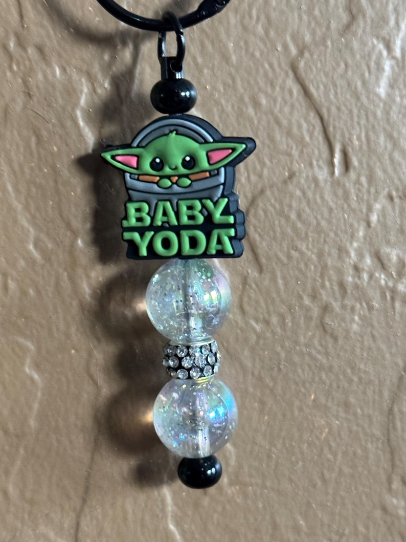 May include: A black and green charm with a cartoon image of Baby Yoda, the words "Baby Yoda" and three iridescent beads.