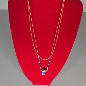 May include: Gold-coloured necklace with a double chain design. The necklace features a pendant with blue and white accents, and small blue and white beads. The text "LEAD COMPLIANT DESIGNED IN USA" is printed on the top.