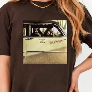 May include: A dark brown t-shirt featuring a graphic of a vintage car with two people inside. The car is light green and has the text "Church Chilling" and "La Bandini's 1970" printed on it. The shirt is worn by a person.