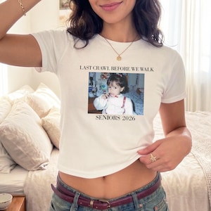 May include: White crop top with the text "LAST CRAWL BEFORE WE WALK" above a small photo of a child. Below the photo is the text "SENIORS 2026". The top is worn by a person.