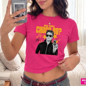 May include: A bright pink short-sleeved t-shirt featuring a graphic print of a person wearing sunglasses and holding a camera. The text above the graphic reads "WHAT WOULD CHUCK DO?" in yellow letters, surrounded by yellow star shapes on the fabric.
