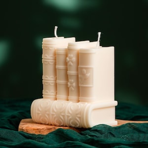May include: Ivory-coloured candle shaped like a stack of books. The candle features detailed book spine designs and visible wicks. The candle sits on a small wooden base against a dark green backdrop.