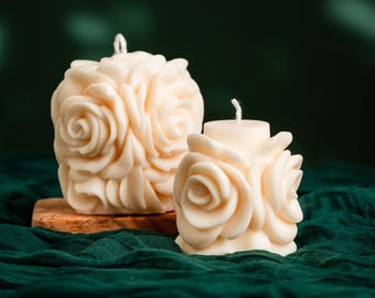 Può includere: Due candele a forma di rosa, color avorio. Una è più grande e poggia su un piccolo blocco di legno, mentre l'altra è più piccola. Entrambe hanno uno stoppino in cima e sono poste su uno sfondo verde scuro.