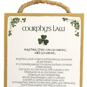 Op de afbeelding: Een wit houten bord met een gouden rand en groene klaverbladontwerpen. Het bord bevat een lijst met humoristische uitspraken over de wet van Murphy, waaronder "Alles wat mis kan gaan, gaat ook mis." en "Murphy was een optimist."