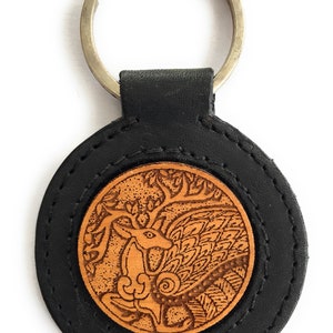 Puede incluir: Un llavero de cuero negro con un dije redondo de cuero marrón que presenta una talla detallada de un ciervo en un árbol.