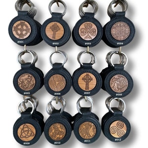 Peut inclure: Une collection de porte-clés en cuir noir avec des motifs celtiques et symboles gravés sur des inserts en bois. Chaque porte-clés est doté d'un anneau métallique et numéroté de #001 à #012.