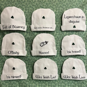 May include: Nine white baby hats with different Irish-themed sayings embroidered on them. The hats have a shamrock design on the top. The sayings include "Leprechaun in disguise", "Full of Blarney", "Irish Roots, American soil", "O'Baby", "Slaintel", "'Tis herself", "'Tis himself", "Wee Irish Lad", and "Wee Irish Lass".