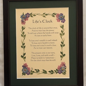 May include: Framed artwork featuring the text "Life's Clock" with a poem about life's fleeting nature. The artwork is framed in a dark brown frame with a green mat. Floral accents in pink, purple, and blue adorn the edges.
