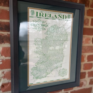 May include: Framed map of Ireland, featuring a detailed green and white cartographic representation of the island. The map is labeled "Clans & Baronies of Ireland" and "Erin Isle". The frame is dark gray, and the background is a brick wall.