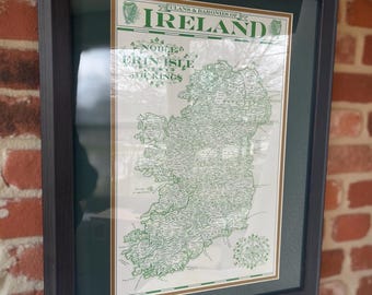 Könnte beinhalten: Gerahmte Karte von Irland mit einer detaillierten grün-weißen kartografischen Darstellung der Insel. Die Karte trägt die Aufschrift "Clans & Baronies of Ireland" und "Erin Isle". Der Rahmen ist dunkelgrau, und der Hintergrund ist eine Ziegelmauer.