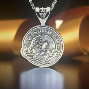Pode incluir: Colar com pingente de prata. O pingente circular tem a inscrição "NON INFERIORA SECUTUS" e um desenho de sol e flor. O pingente pende de uma delicada corrente de prata.
