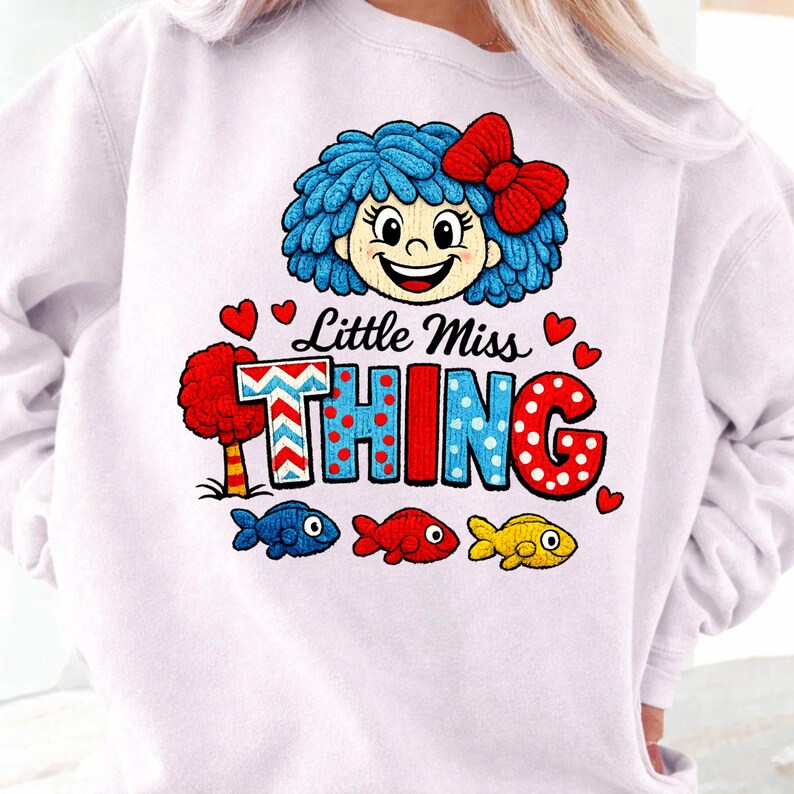 May include: White sweatshirt with a cartoon graphic of a girl with blue hair and a red bow. The text "Little Miss Thing" is displayed with colourful letters, alongside heart and fish graphics.