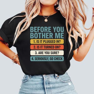 Könnte beinhalten: Schwarzes T-Shirt mit dem Text "BEFORE YOU BOTHER ME" in Türkis. Darunter listen vier Zeilen Fragen in weißem Text auf farbigen Balken auf: gelb, orange, rot und türkis. Das T-Shirt wird von einer Person getragen.
