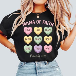 Puede incluir: Camiseta negra con el texto rosa "MAMA OF FAITH". Debajo, nueve caramelos en forma de corazón de varios colores, cada uno con una palabra como "LOVE" o "JOY". El texto "Proverbios 31:28" está en la parte inferior.