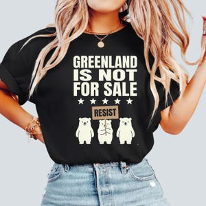 Może przedstawiać: Czarny t-shirt z białym napisem "GREENLAND IS NOT FOR SALE" i pięcioma gwiazdami. Poniżej trzy białe ilustracje niedźwiedzi polarnych, z których jeden trzyma znak z napisem "RESIST".